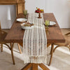 A wooden dining table set for a meal with a white crocheted tasseled table runner and rustic tableware.