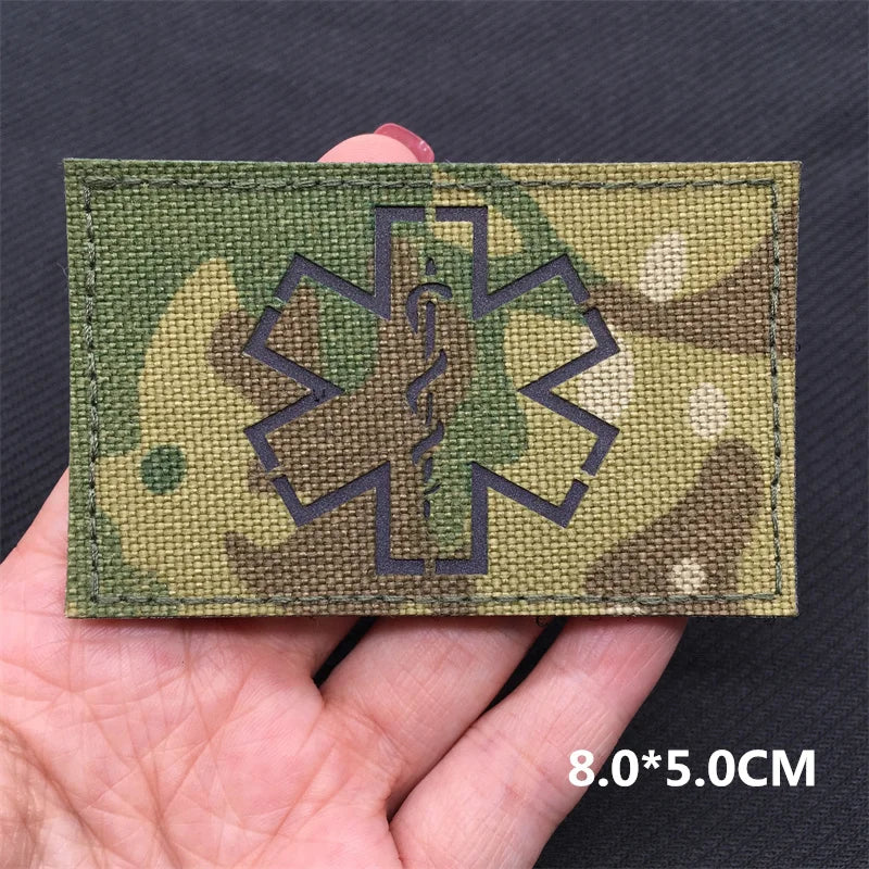 A rectangular tactical patch featuring a dark silhouette of a medical cross on a camouflage background being held by a hand.