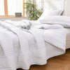 White quilted summer blanket folded on a bed with pillows and a window in the background