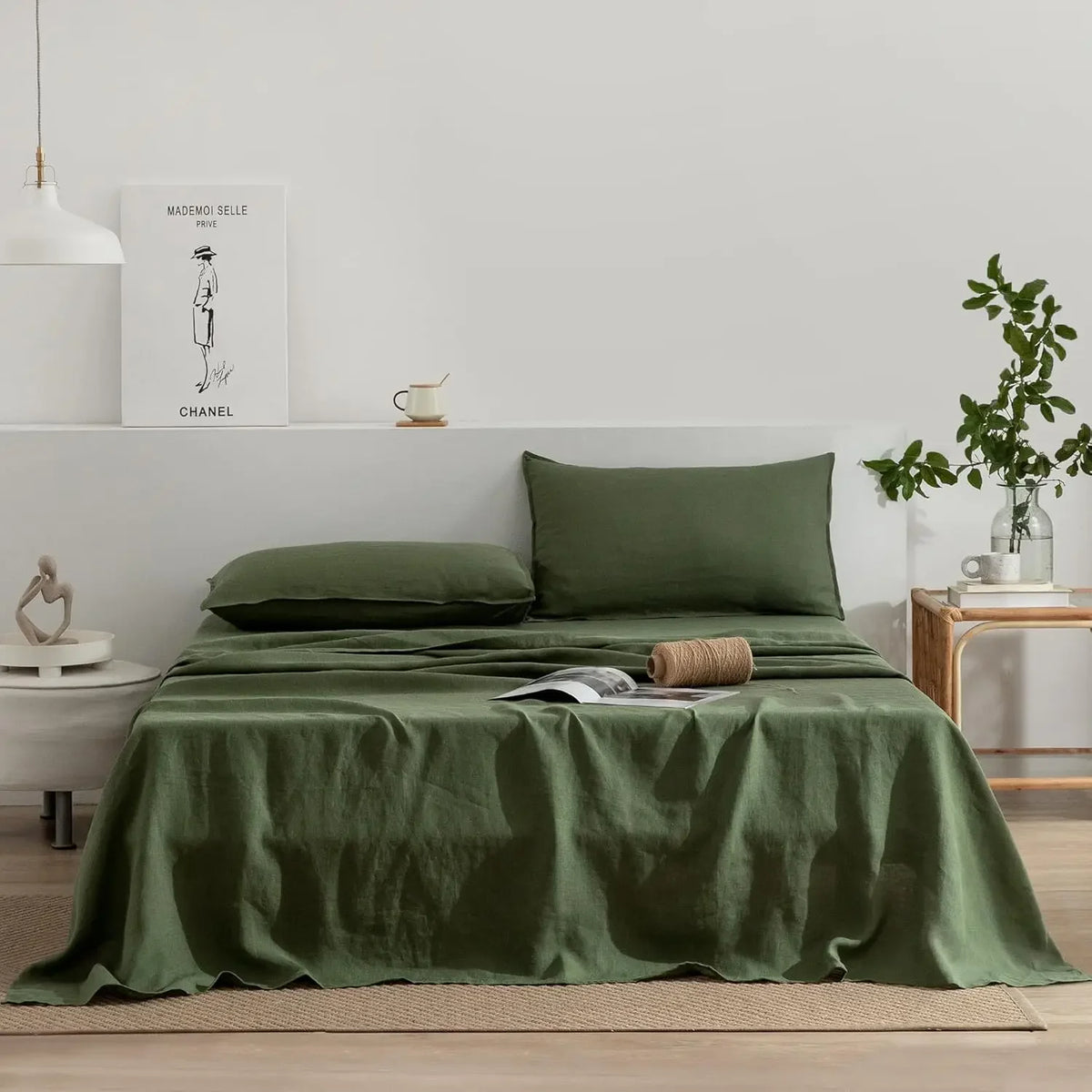 A green pure French linen bed sheet is spread on a bed with a matching pillow and a framed Chanel print in the background.