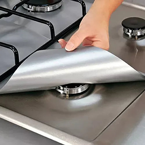A hand is placing a silver reusable gas hob protector liner onto a stovetop burner.