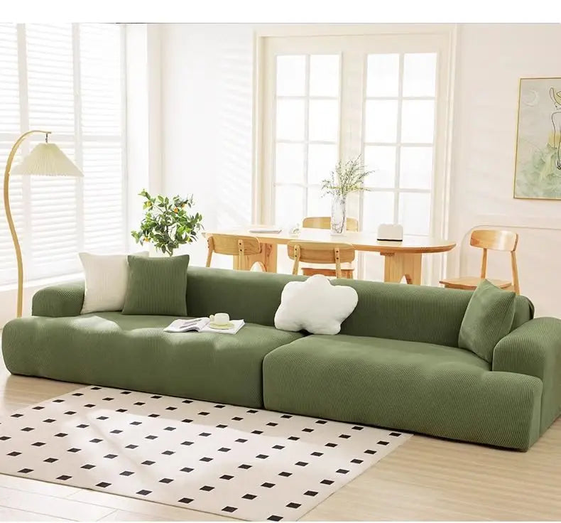 A modern, olive green stretchable sofa slipcover with white cushions and a black and white patterned rug in a bright room.