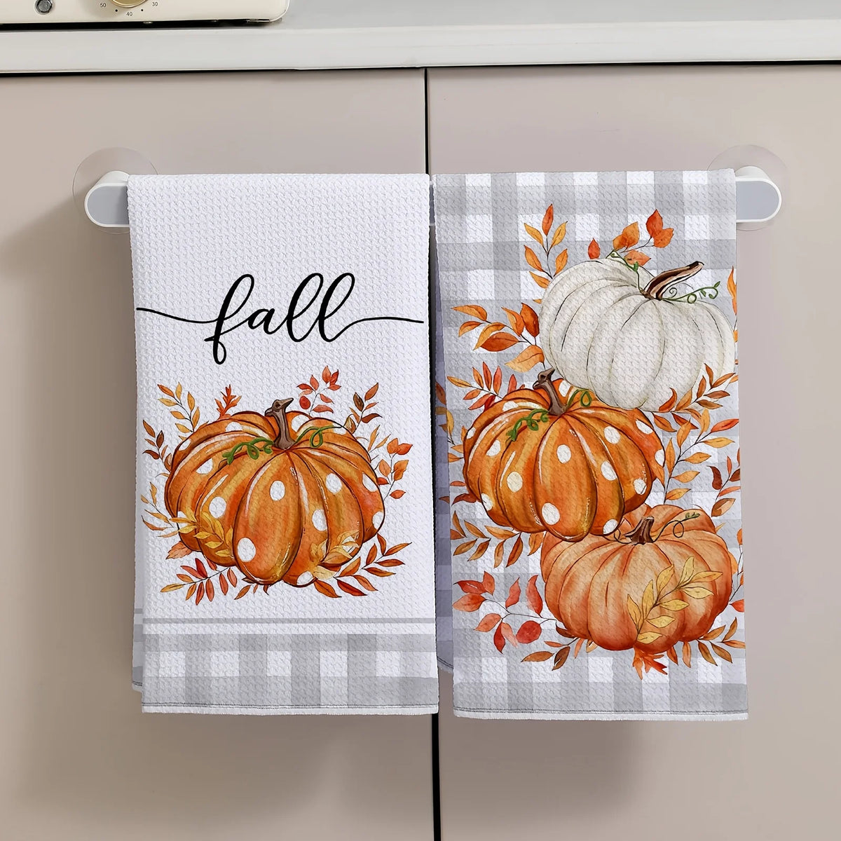 A white kitchen towel featuring an orange pumpkin print with autumn leaves and the word fall.