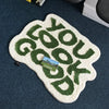 A white foot mat with green embossed text that reads 'GOOD YOU GOOD' is placed on a dark blue surface.