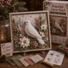 A framed piece of fabric art featuring white and pink floral and evergreen elements with a wooden tool.