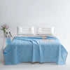 A light blue quilted blanket is spread on a bed with two white pillows and a small silver clock.
