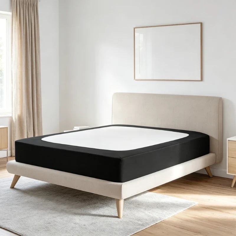 A white box spring protector cover is displayed on a beige bed frame with a black mattress, set against a white wall.