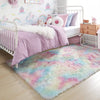 A fluffy rainbow colored area rug in pink, blue, and white is placed on a wooden floor next to a white crib and storage cabin