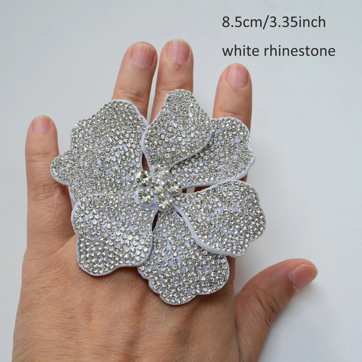 A hand holds a white rhinestone flower applique patch, measuring 8.5cm or 3.35 inches.