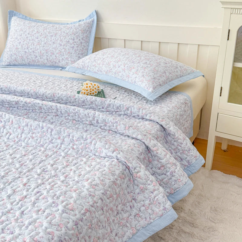 A light pink and blue floral quilted bedspread is displayed on a bed with white wooden headboard.