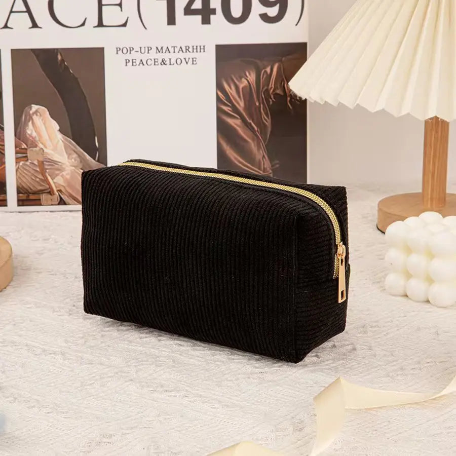 Black ribbed cosmetic storage pouch with a gold zipper resting on a white textured surface.