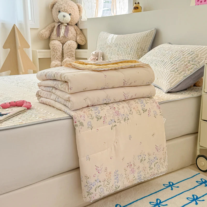 A stack of light beige quilted blankets and pillows on a white surface with a teddy bear in the background.