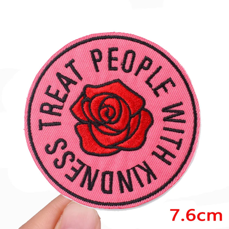 A round pink embroidered patch with the text Treat People With Kindness and a red rose design.