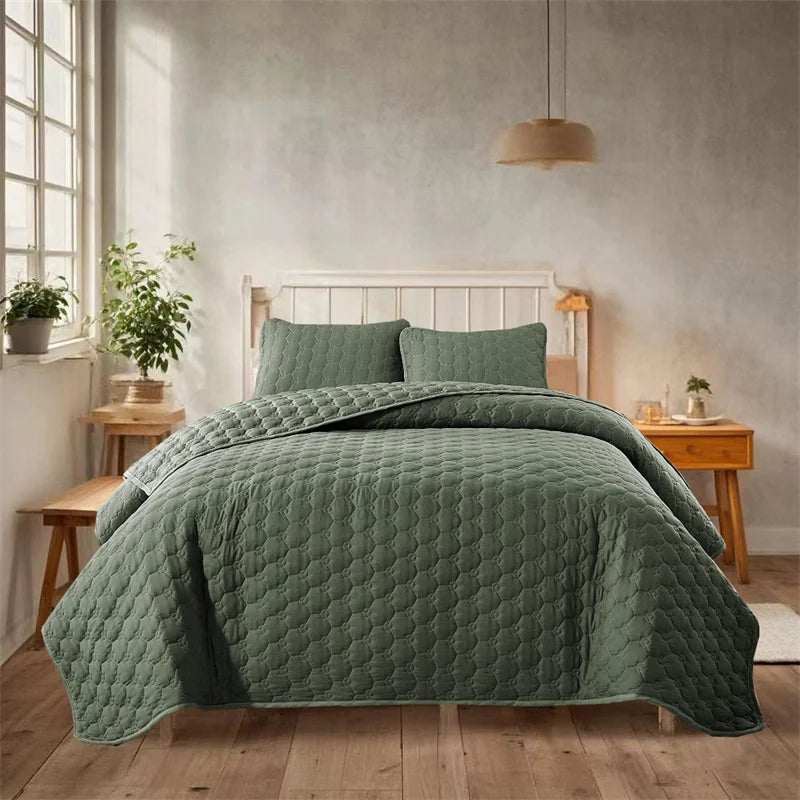 A teal quilted bedspread set with green accents is displayed on a bed in a bright, minimalist bedroom.
