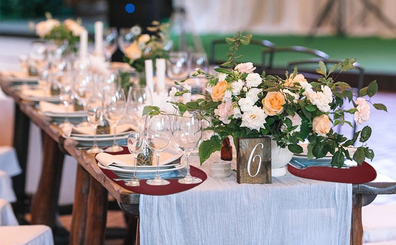 A wooden dining table set with glassware, white plates, and a floral centerpiece on a light blue tablecloth.