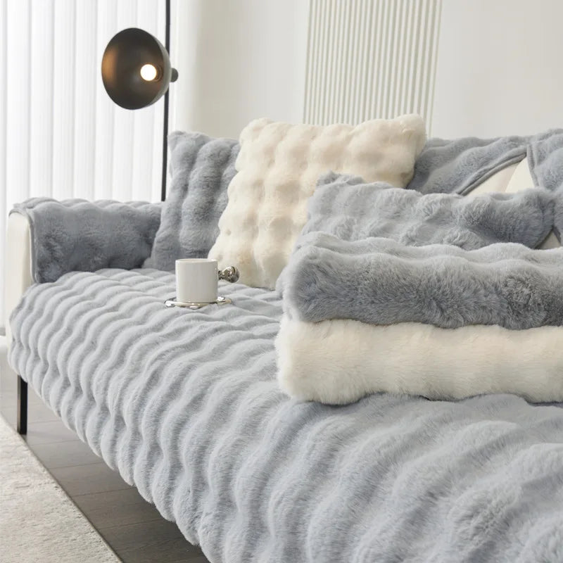 A gray plush velvet sofa cover with white and gray faux fur pillows on a bed.