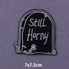 Black embroidered patch shaped like a tombstone with the text Still Horny and a small flower detail.