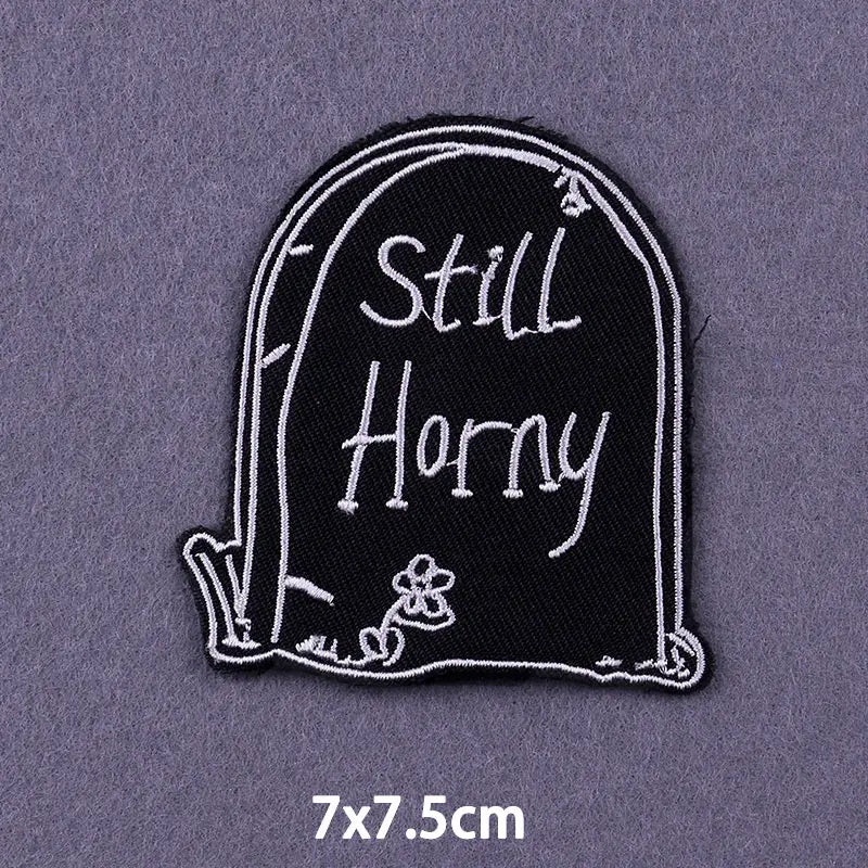Black embroidered patch shaped like a tombstone with the text Still Horny and a small flower detail.