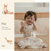A baby wearing a white cotton sleeping bag with a fox pattern is lying on a brown blanket.