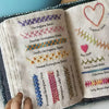 A close-up of a beginner embroidery stitch book showing various herringbone and other decorative stitches on cream fabric.