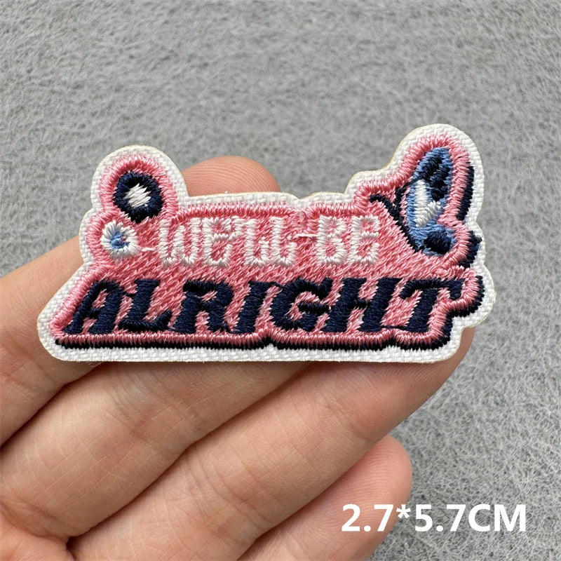 A hand holds a pink embroidered patch with the text WELL BE ALRIGHT in dark blue and white thread.