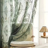 A sheer lace floral window curtain in sage green and white hangs beside a light-colored upholstered chair.