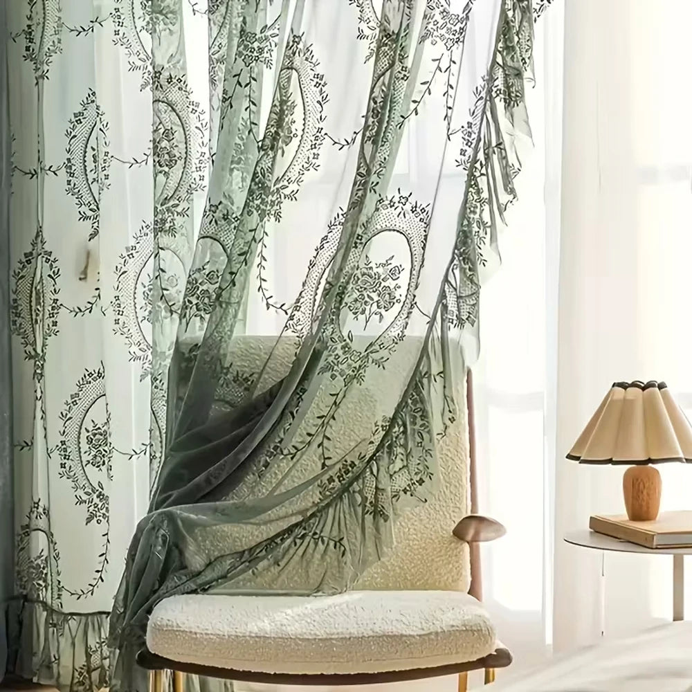 A sheer lace floral window curtain in sage green and white hangs beside a light-colored upholstered chair.