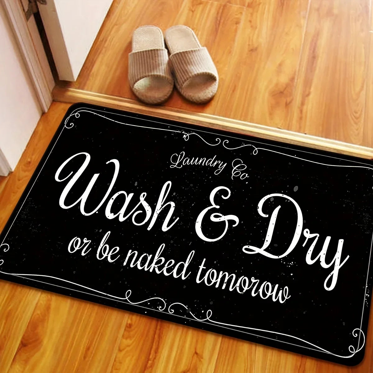 A black laundry mat with white text that reads Wash & Dry or be naked tomorrow, placed on a wooden floor.