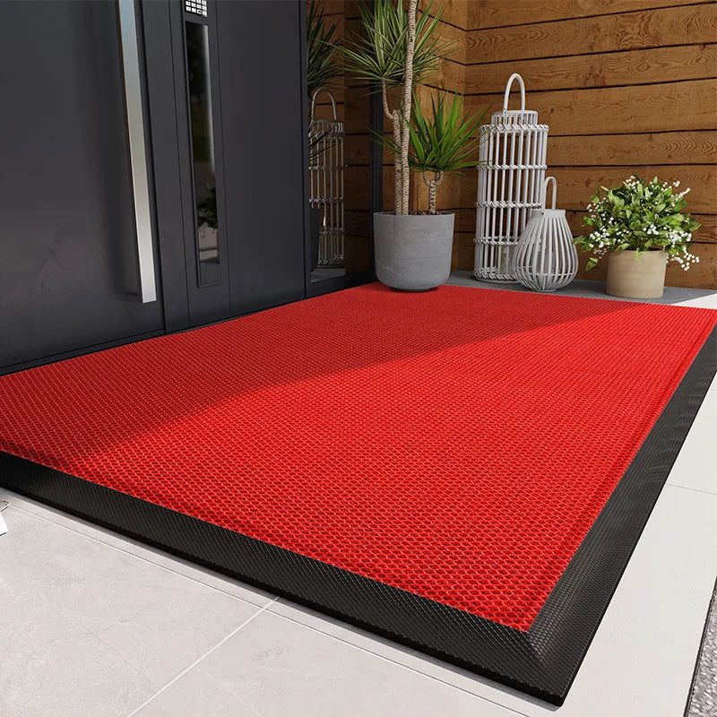 A bright red non-slip rubber doormat placed on a light gray floor near a wooden door with potted plants.