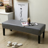 A gray dining bench cover is displayed on a wooden bench, with a drawing of a woman on the wall and some slippers nearby.