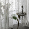 Sheer floral lace window curtains hanging in a bright room with a wooden table and a classical statue.