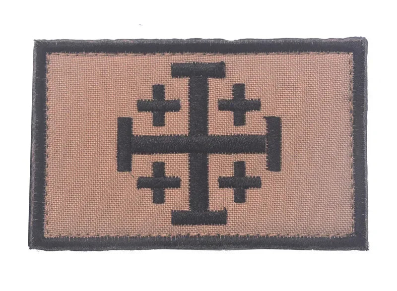 A rectangular embroidered patch featuring a black cross design on a tan fabric background.