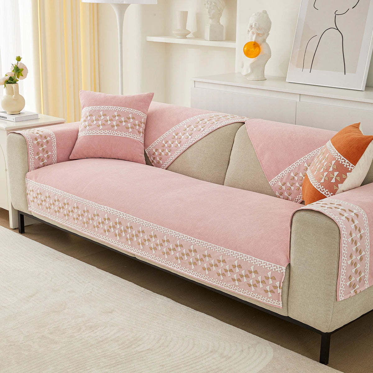 A pink quilted sofa cover with lace trim and beige fabric is displayed on a sofa in a bright living room.