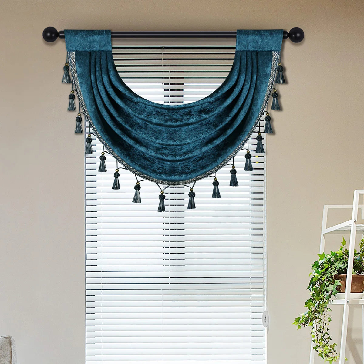 A dark teal velvet window curtain valance with fringe tassels is displayed above a white horizontal blind.