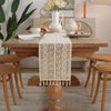 A vintage crochet lace table runner with fringe is displayed on a wooden dining table surrounded by natural light and greener