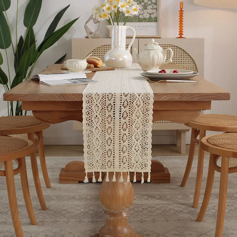 A vintage crochet lace table runner with fringe is displayed on a wooden dining table surrounded by natural light and greener