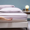 A hand gently touches a white duvet on a bed with light purple pillows and a bedside lamp.