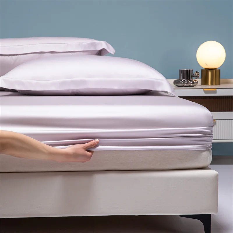 A hand gently touches a white duvet on a bed with light purple pillows and a bedside lamp.