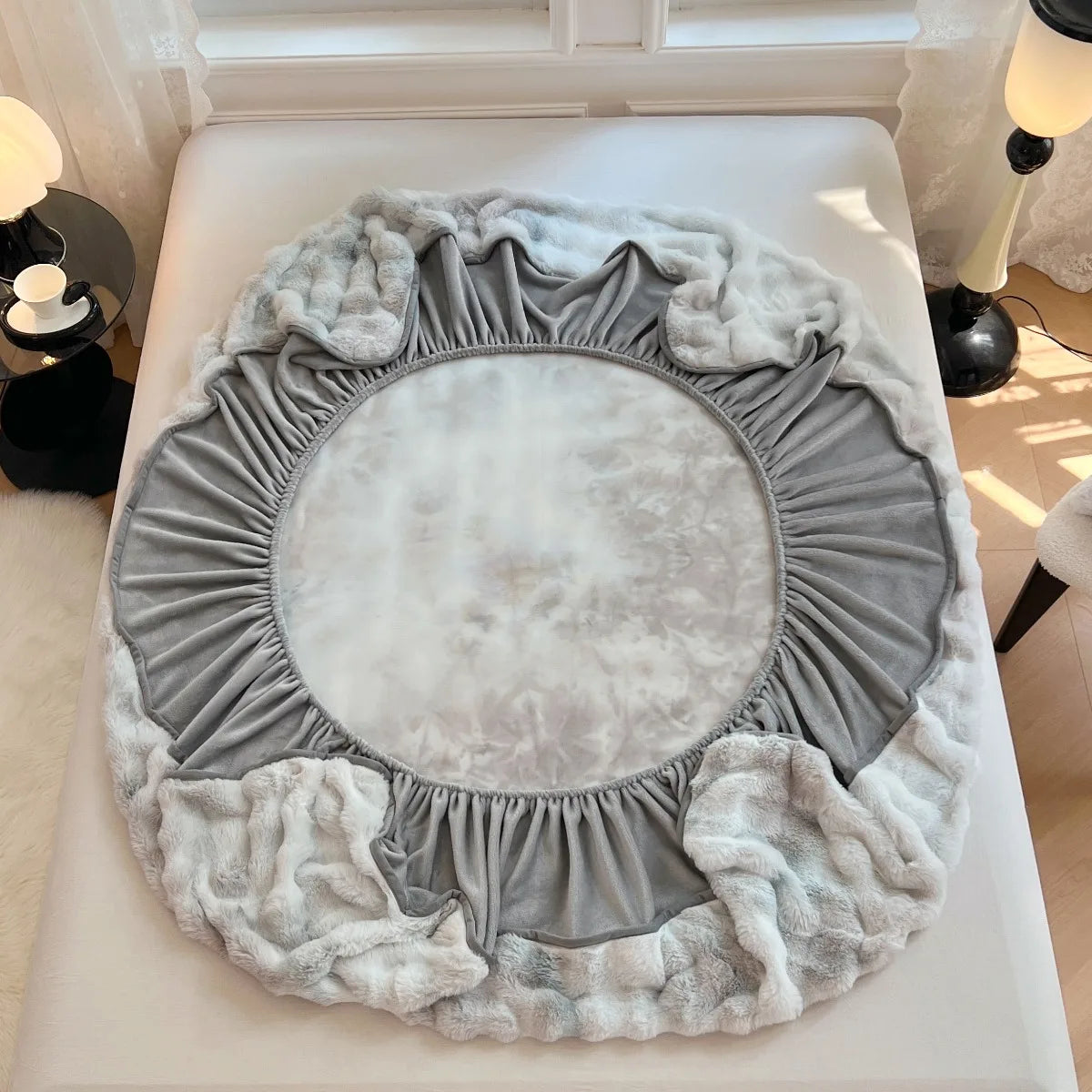 A round, ruffled faux rabbit fur blanket in gray and white rests on a white surface.