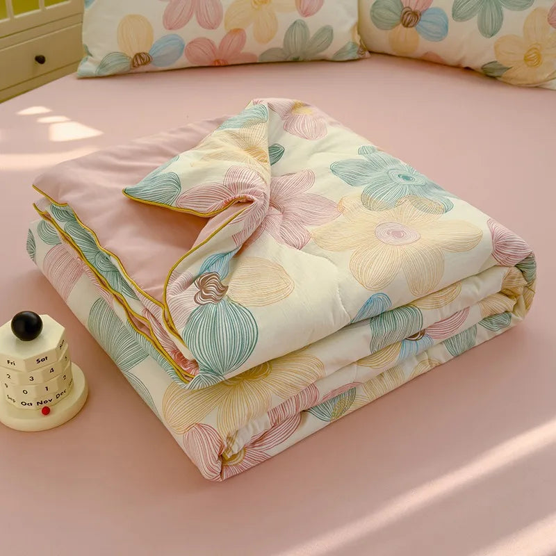 A folded Summer Gauze Cotton quilt with floral patterns in pink, yellow, and blue tones resting on a pink surface.