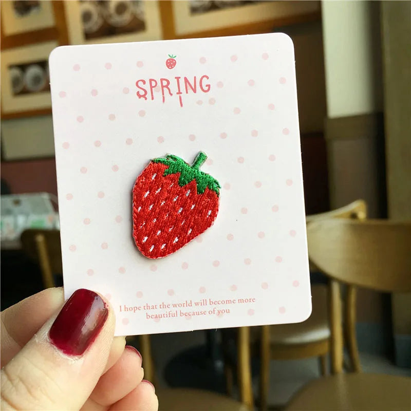 A hand holds a white card featuring a red strawberry embroidery patch and the word SPRING.