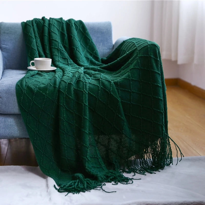 A deep emerald green knitted tassel throw blanket draped over a blue sofa with a white cup nearby.