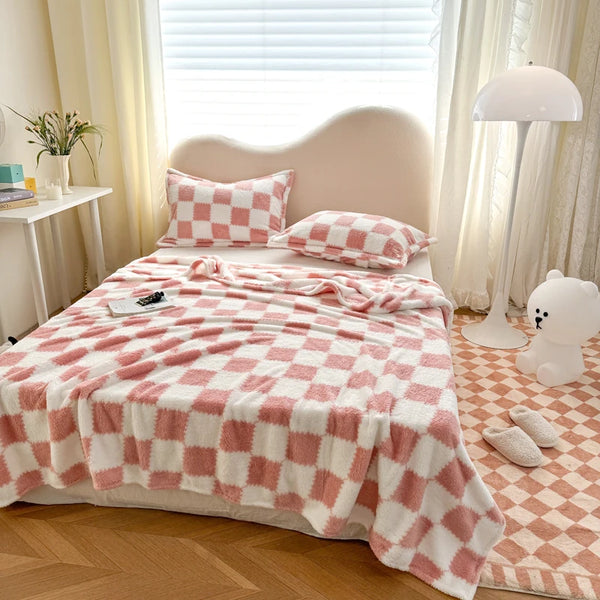 A pink and white checkered flannel blanket is displayed on a bed with a headboard, next to a lamp.