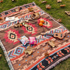 A woven blanket with geometric patterns in earth tones laid out on green grass, featuring dried leaves and a wooden guitar.