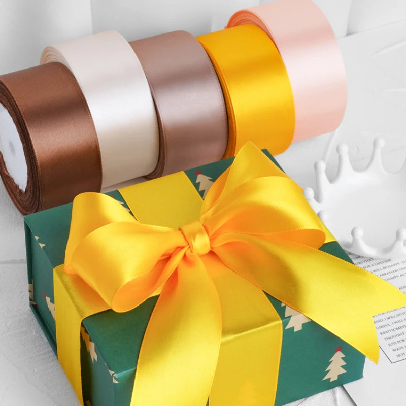 A gift box wrapped with a yellow satin ribbon and a roll of multi-colored decorative ribbons.