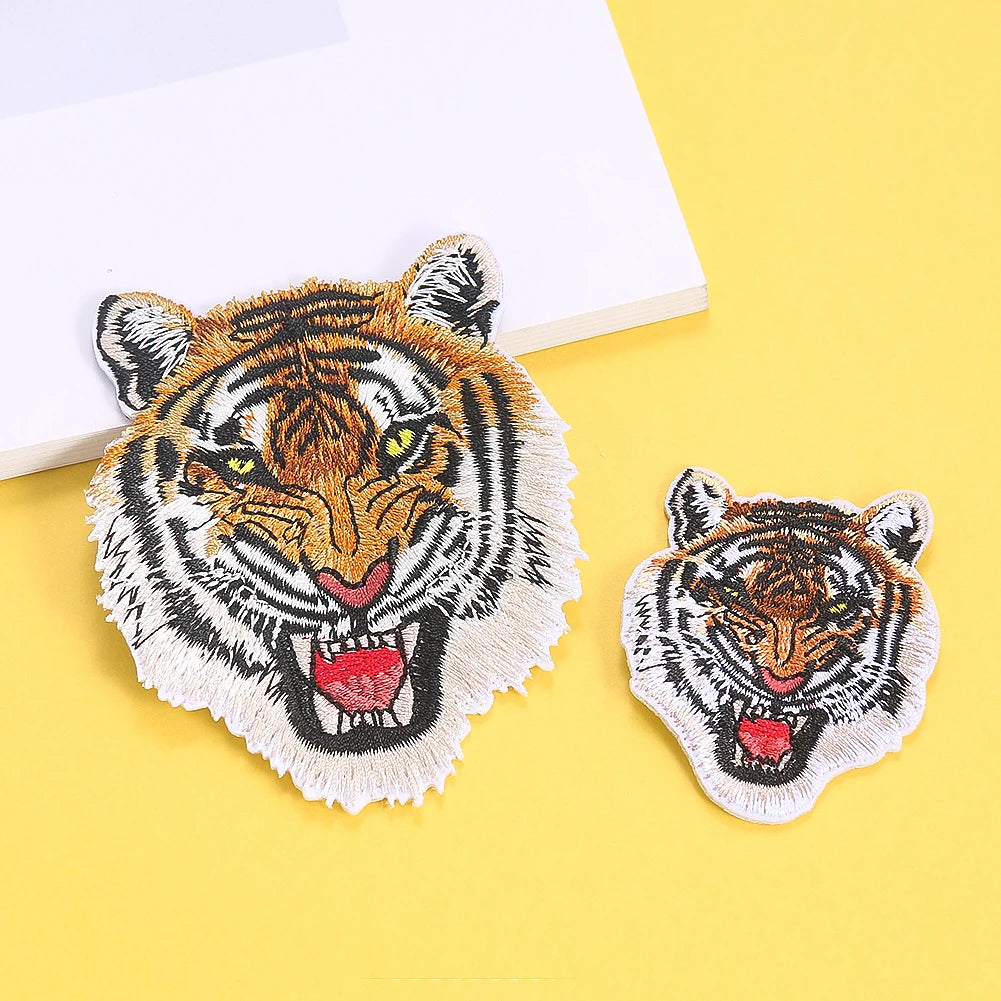 A detailed embroidery patch of a tiger's face with yellow eyes and open mouth on a light background.