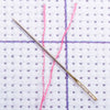 A thin gold needle is positioned across a white fabric background with purple and pink threads.