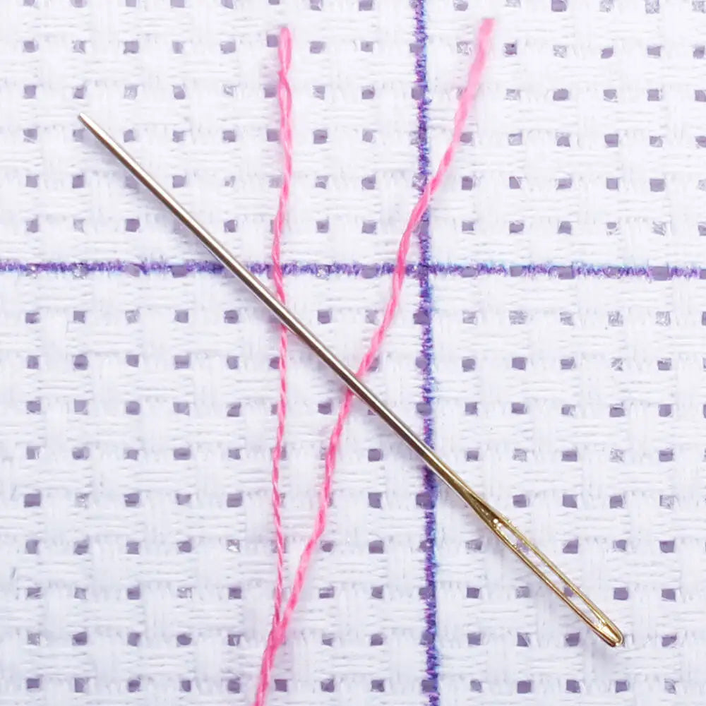 A thin gold needle is positioned across a white fabric background with purple and pink threads.