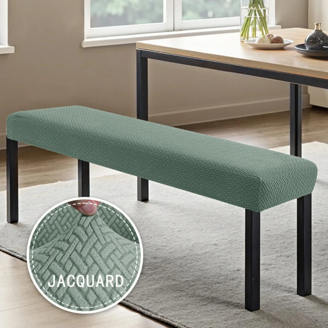 A green stretch jacquard dining bench cover placed on a wooden table with a glass vase in a bright room.