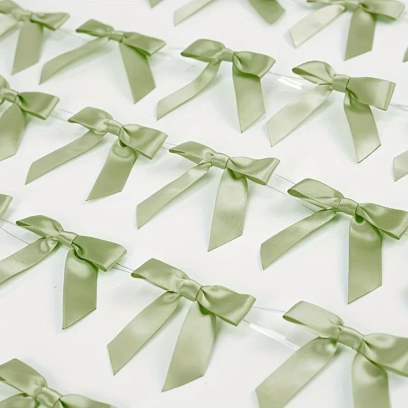 A close-up of a 50 pack of sage green satin ribbon twist tie bows arranged in a line.